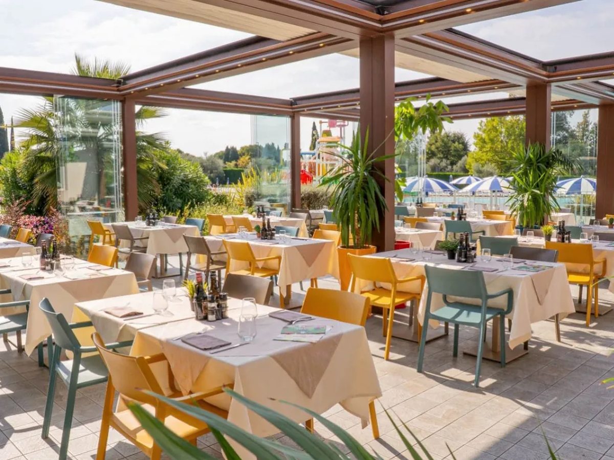A covered restaurant terrace under a timber and glass roof, with tables in cream cloths set with glasses, yellow and green chairs, and gardens and blue-and-white pool parasols beyond the glazing.