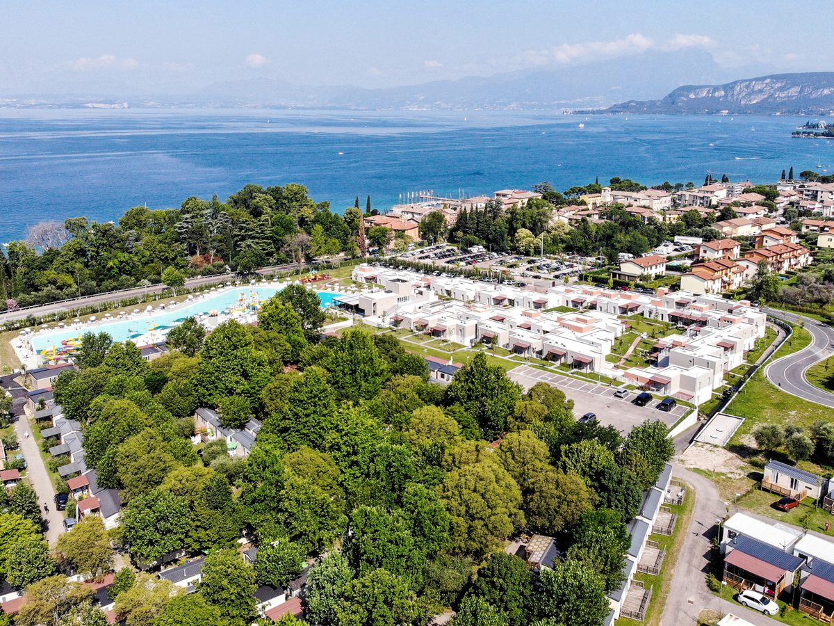 Aerial view of Sisan Family Resort: rows of white flat-roofed apartment blocks and a lagoon pool with coloured slides, the wooded campsite and the lakeside road in front, and the deep blue expanse of Lake Garda beyond.