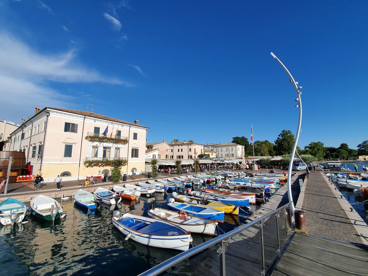 A Lake Garda harbour lined with colourful wooden rowing boats and small motorboats at their moorings, a pale yellow town hall and waterfront cafes behind, and a curved steel lamp standard on the quay.