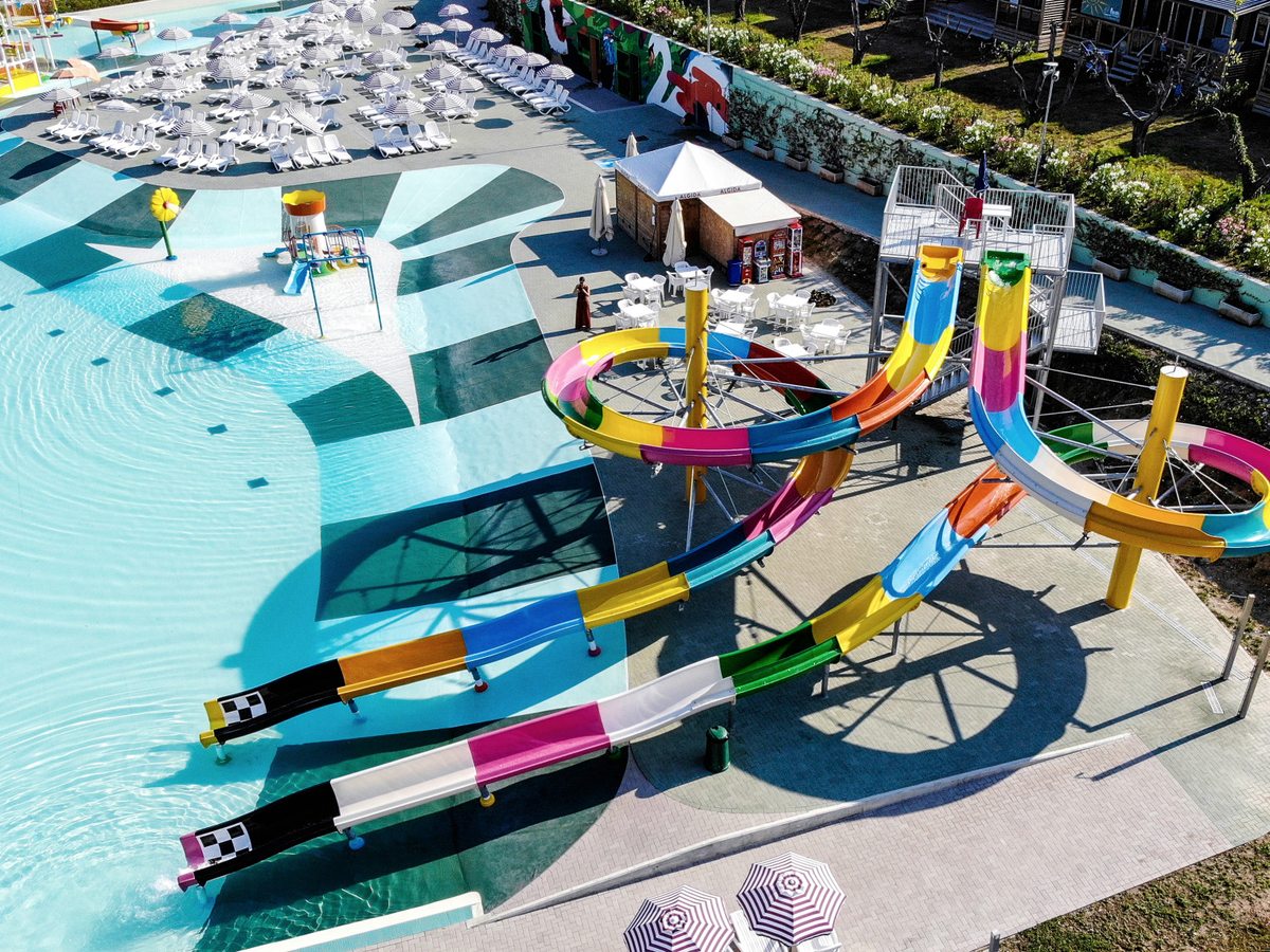 Coloured spiral flume slides and multi-lane racing slides running down into the shallow edge of the lagoon pool, with rows of white loungers and striped parasols on the terrace beyond.