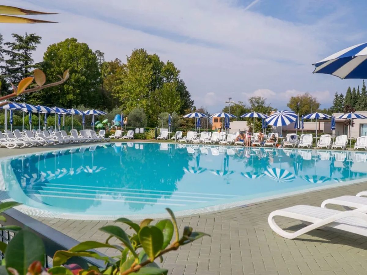 A large swimming pool with pale blue water, ringed by white sun loungers and blue-and-white striped parasols, with lawn and tall mature trees behind.