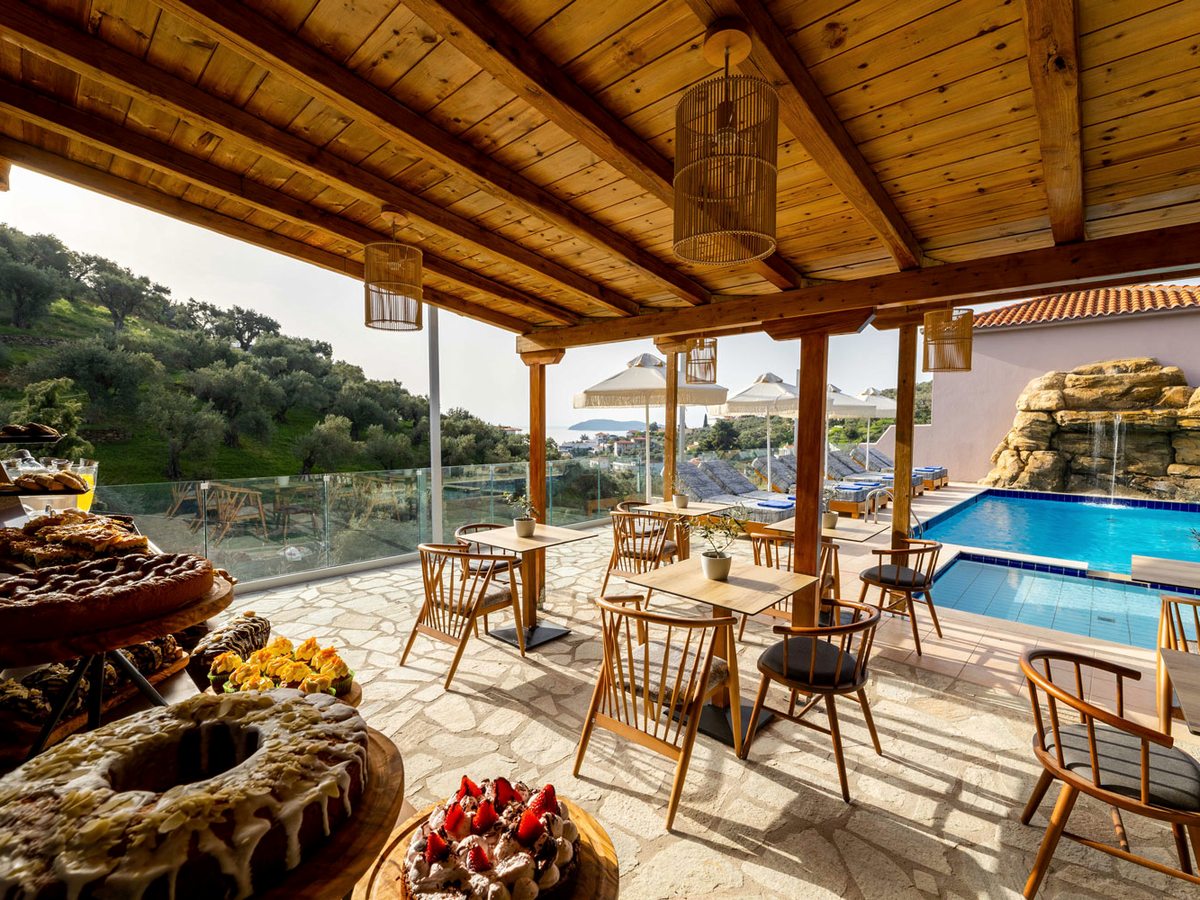 A covered terrace with a timber roof and woven lanterns, wooden tables and chairs on a stone floor, cakes and pastries on a counter in the foreground, and the pool and an olive-covered hillside beyond.