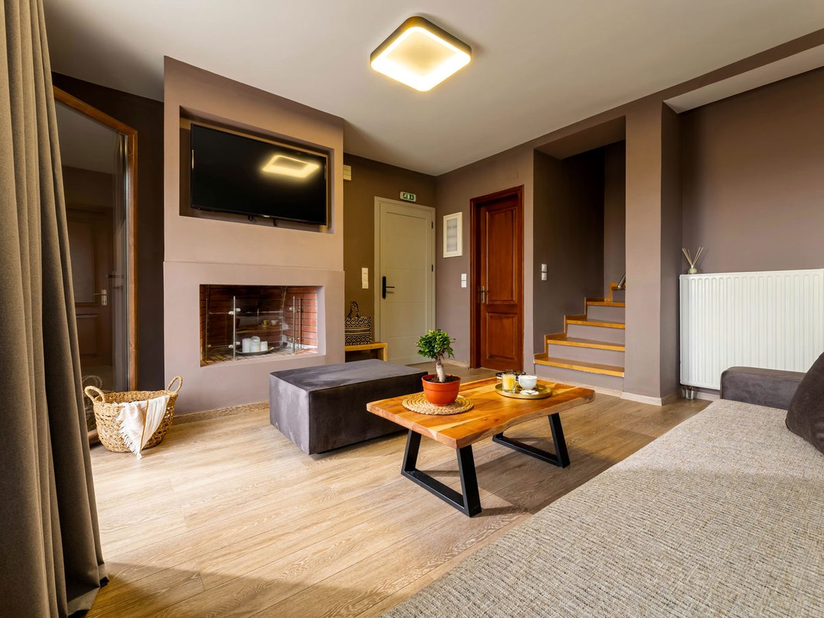 A living room with a built-in fireplace below a wall-mounted television, a wooden coffee table and grey ottoman on a light wood floor, and a staircase to one side.