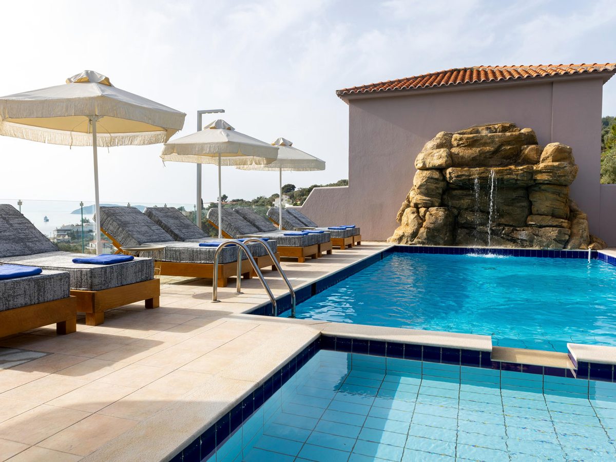 A shallow tiled pool stepping down in the foreground beside the main pool, with sun loungers under fringed parasols and the rock waterfall behind.
