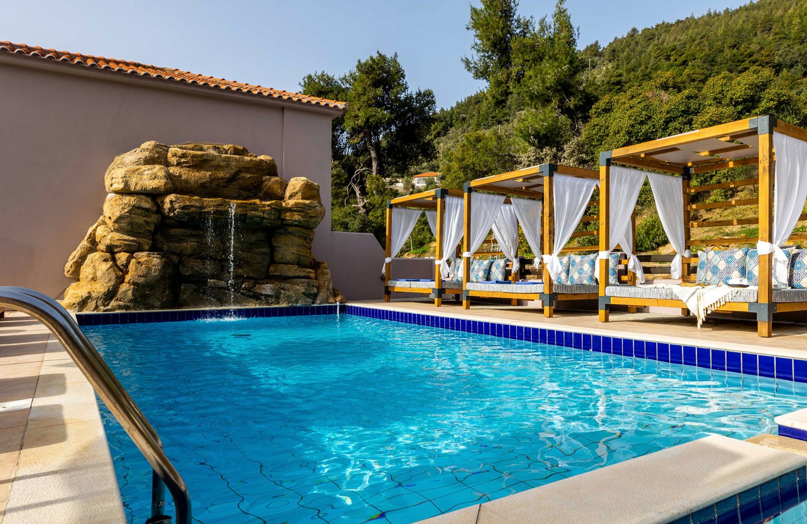 An outdoor pool with a rock waterfall at one end and curtained wooden daybeds along the far side, backed by a pine-covered hillside.