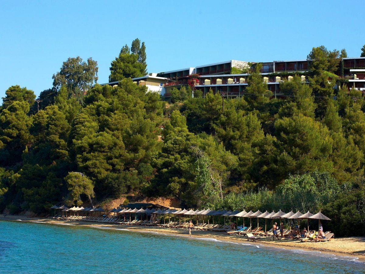 The long hotel buildings on a pine-covered hillside above a beach lined with thatched parasols and wooden sunbeds, with clear shallow water in front.