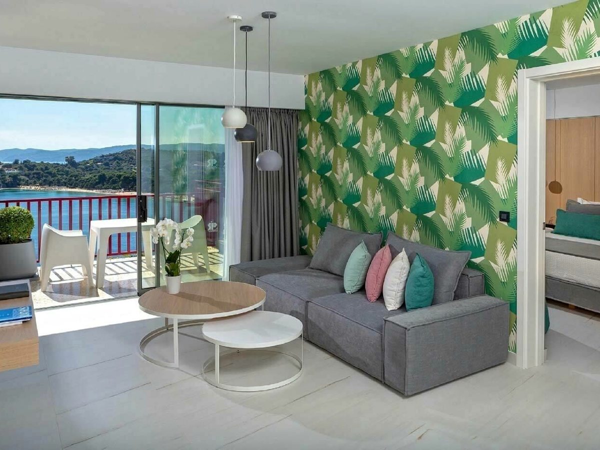 A suite living room with a grey corner sofa against palm-print wallpaper, nesting coffee tables, glass doors to a balcony and a bedroom through an open doorway.