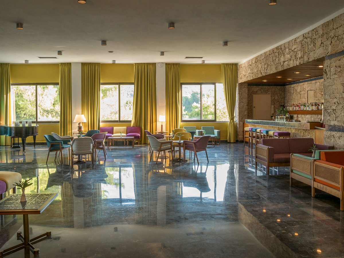 An indoor lounge with a bar counter along a stone wall, armchairs and low tables on a marble floor, tall curtained windows and a grand piano.