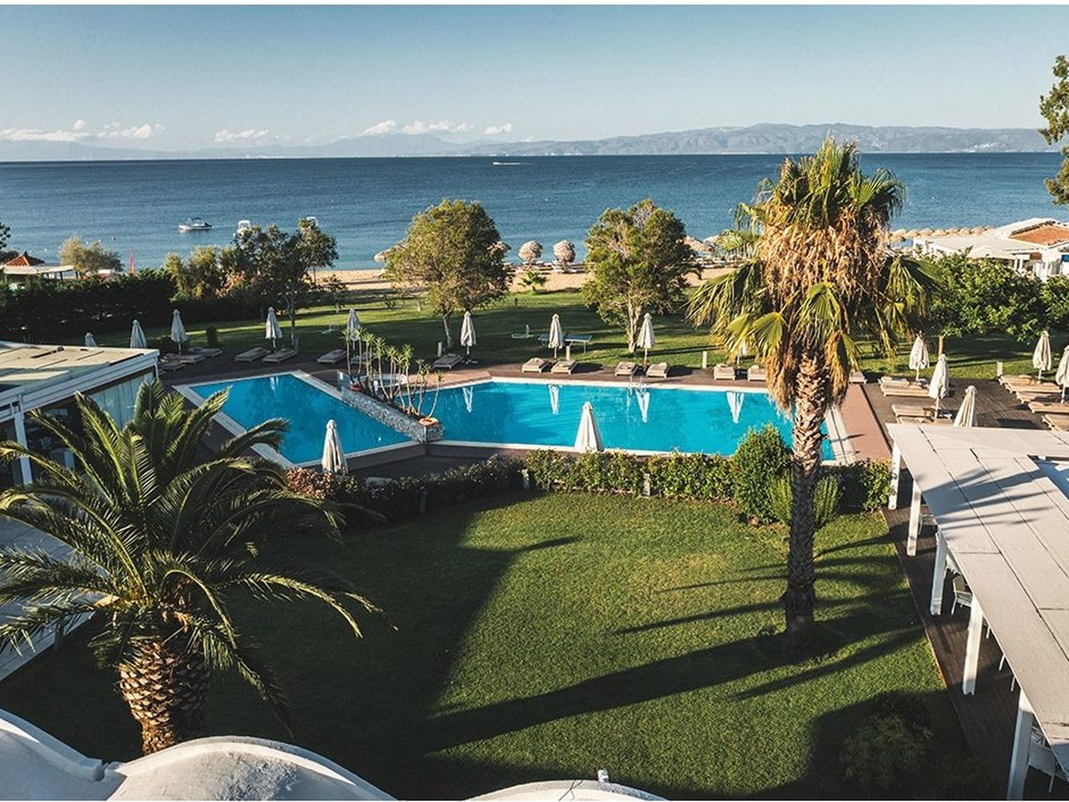 Two blue pools edged with sun loungers and furled parasols in a lawned garden with tall palms, with a sandy beach lined with thatched umbrellas and the open sea and distant mountains beyond.