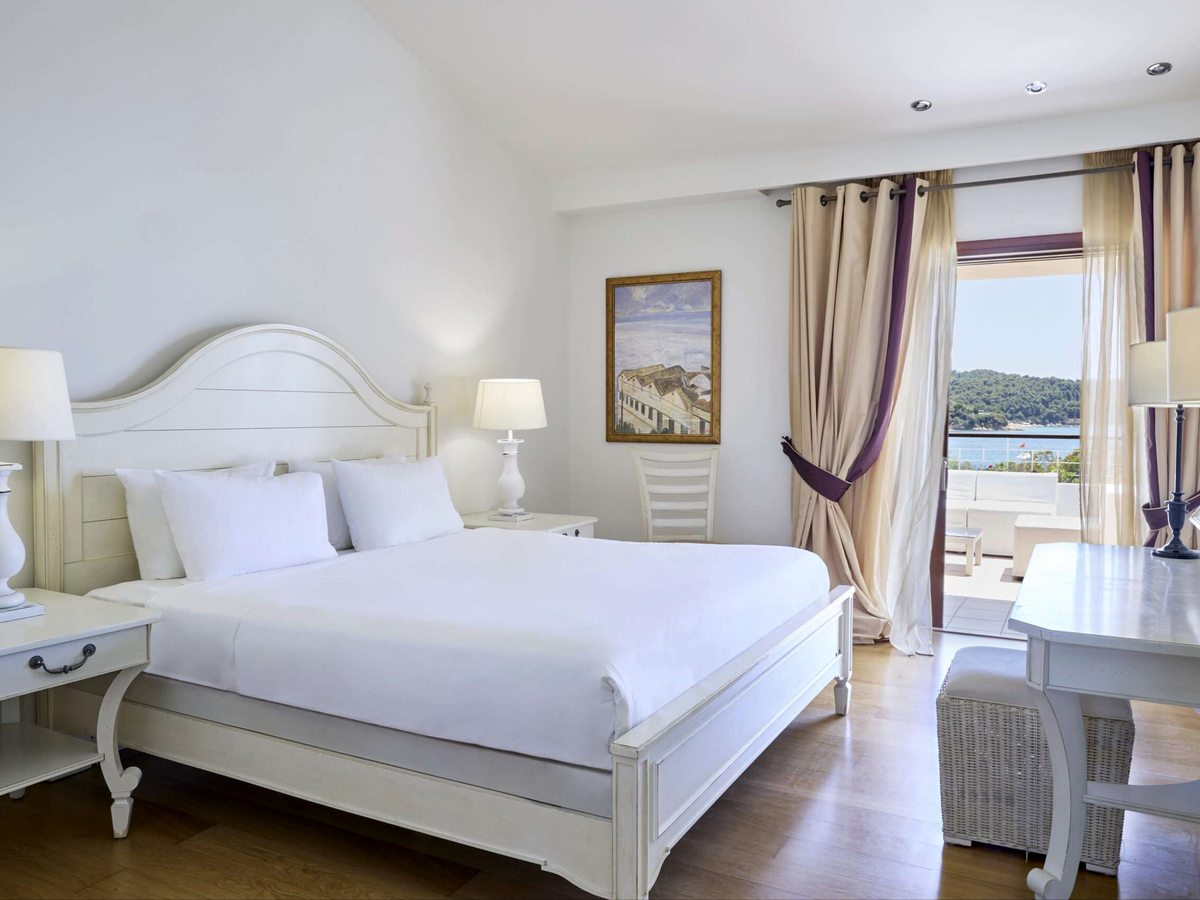A double bed with a white painted headboard and white bedding, matching bedside tables and lamps, a framed seascape and open doors to a balcony looking over the wooded bay.
