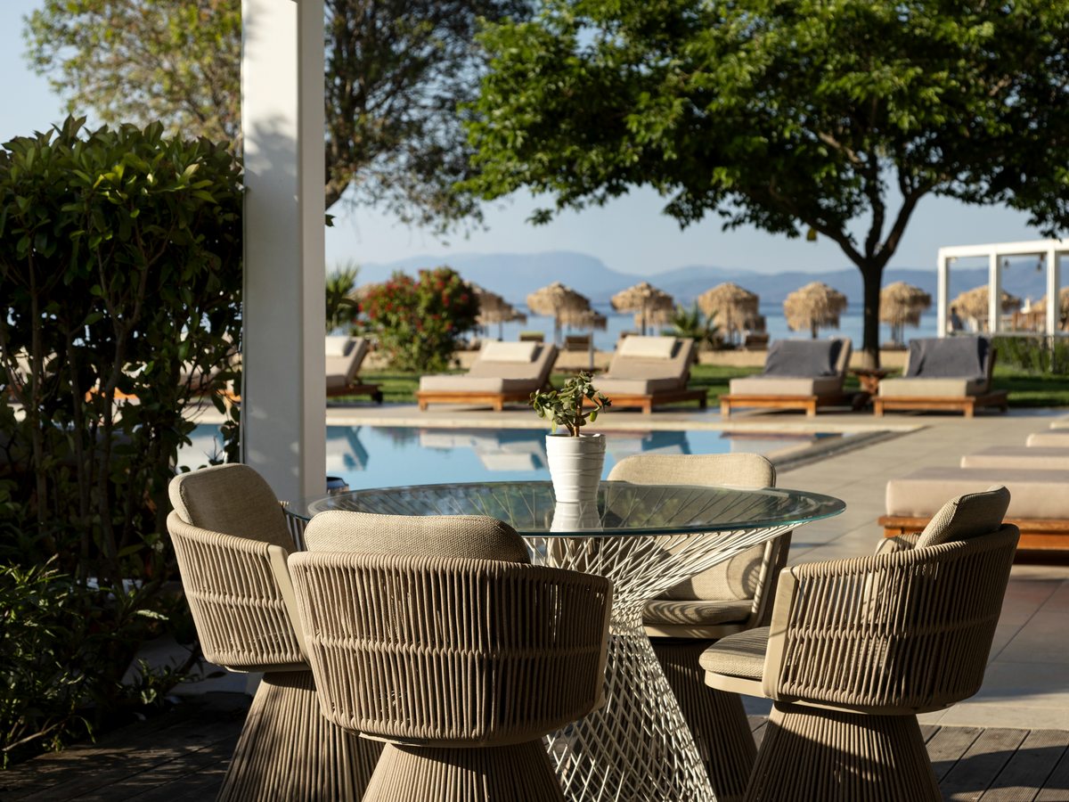 A round glass table and four woven armchairs on a shaded terrace beside a pool, with sun loungers, thatched beach umbrellas, the sea and distant mountains beyond.