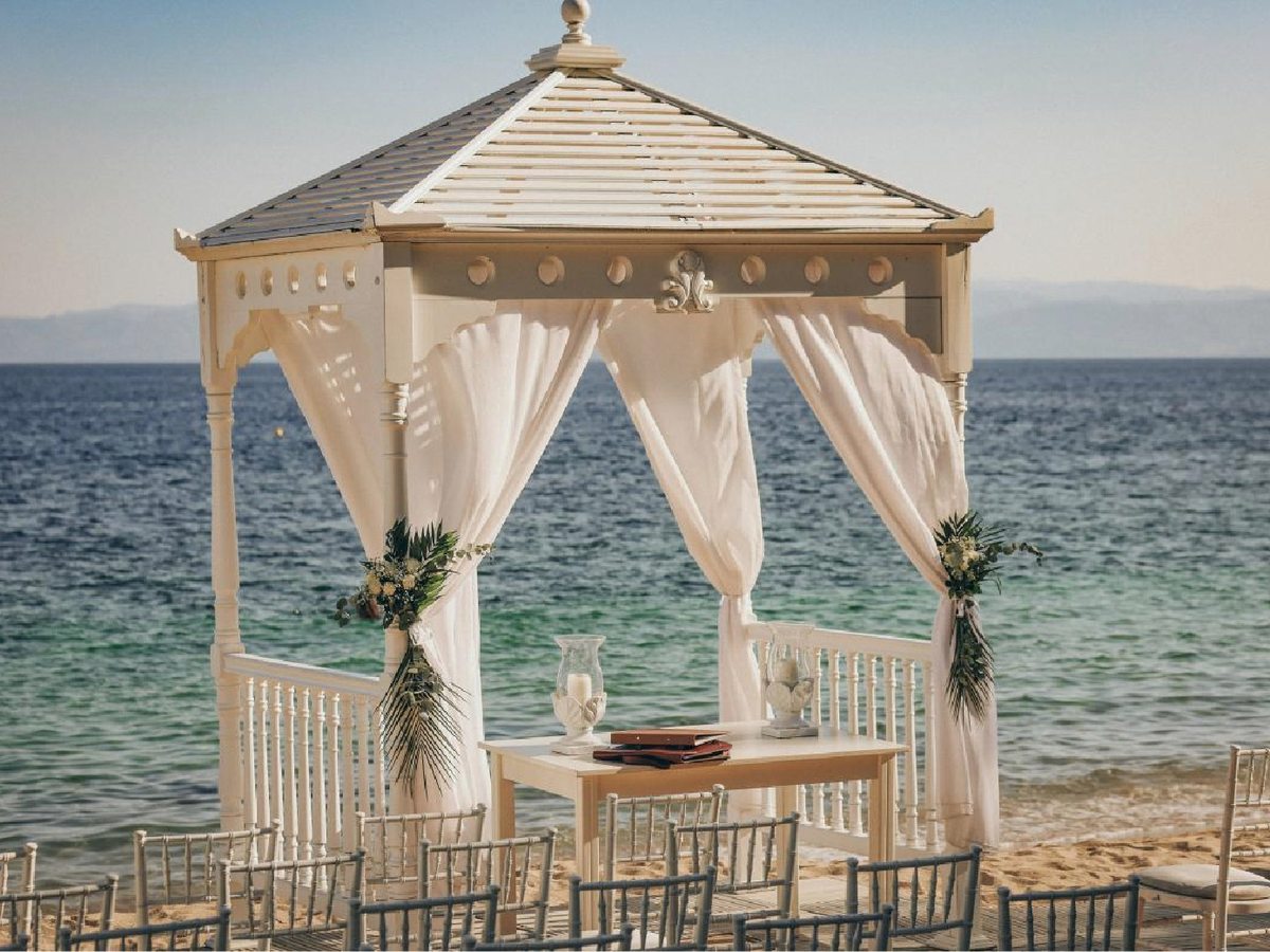 A white timber gazebo with curtained sides and floral posies standing on the sand at the water's edge, a small table inside and rows of chairs in front.