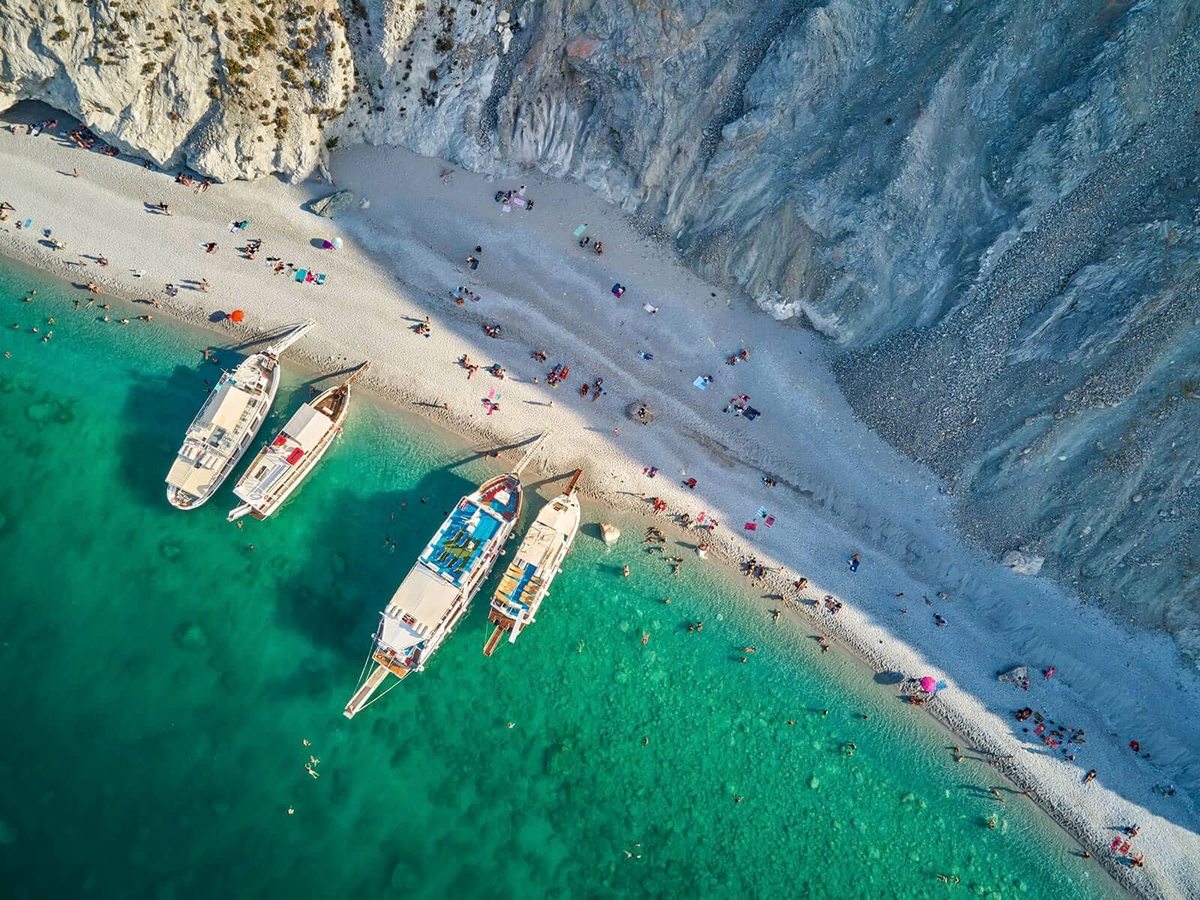A narrow white pebble beach at the foot of pale cliffs, with turquoise water, sunbathers and four wooden excursion boats moored at the shore.