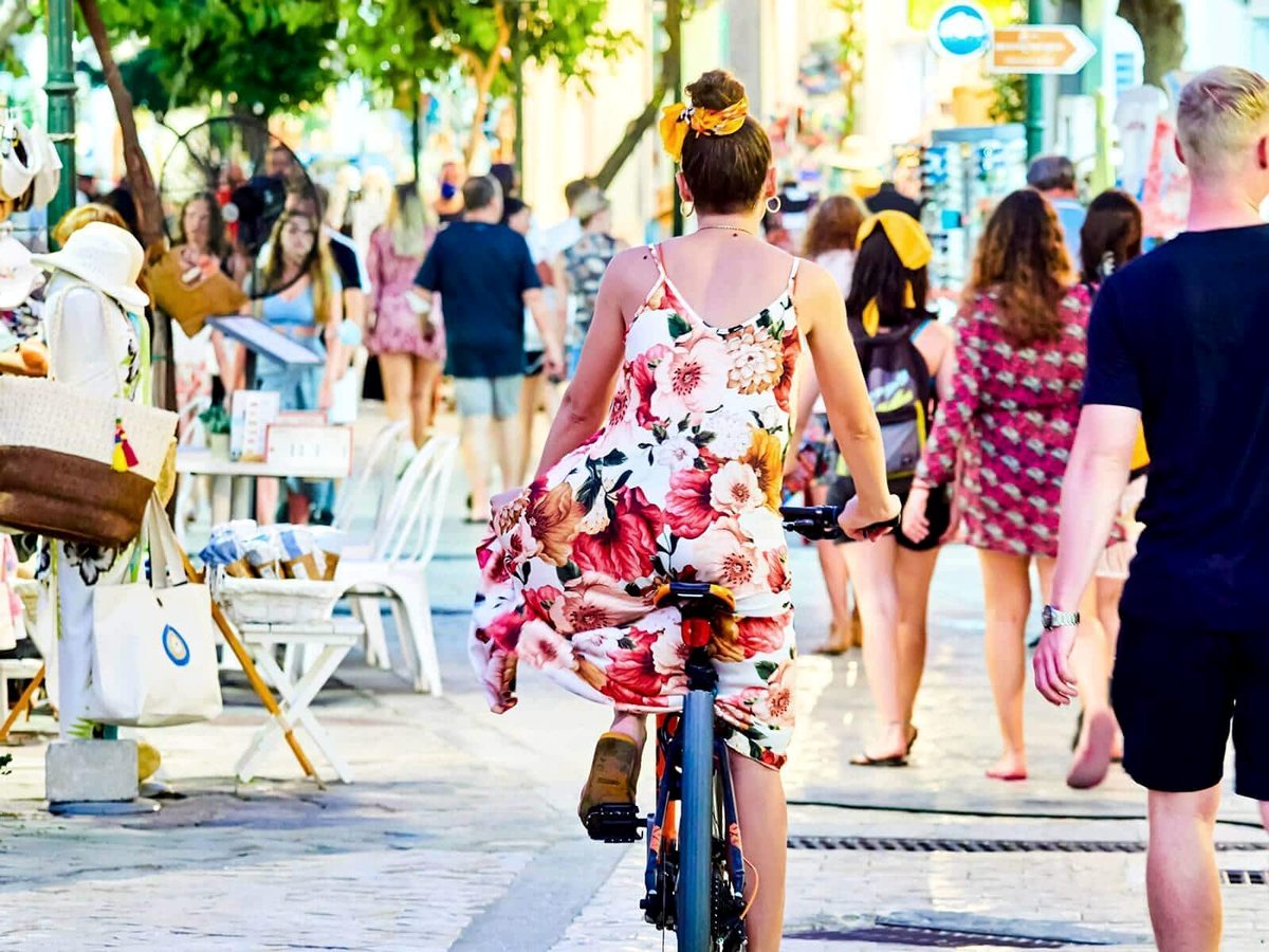 A busy pedestrian shopping street with a woman cycling, market stalls of bags and hats, and shoppers walking under trees.