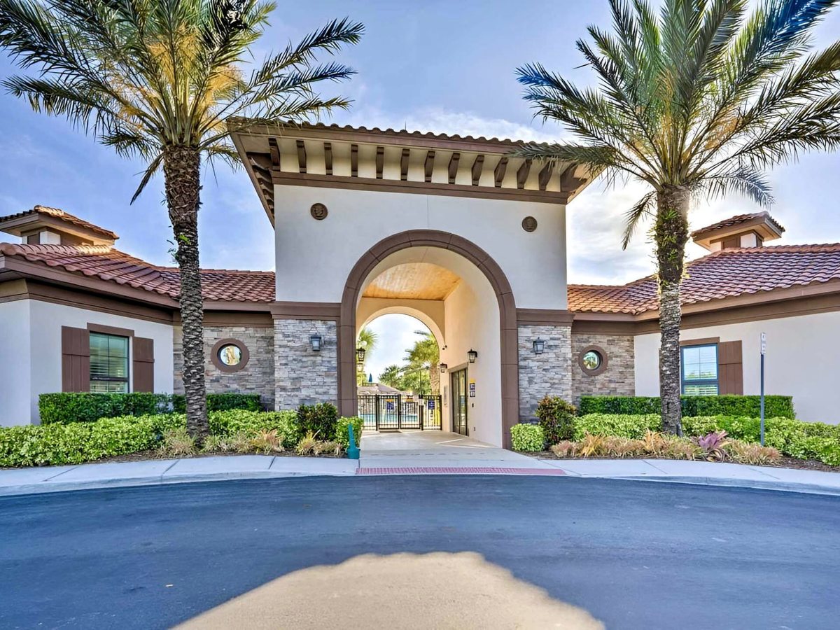 A wide cream Mediterranean-style building with a tall central archway, stone-clad columns, a terracotta-tiled roof and tall palm trees, seen across an empty tarmac drive under a blue sky.