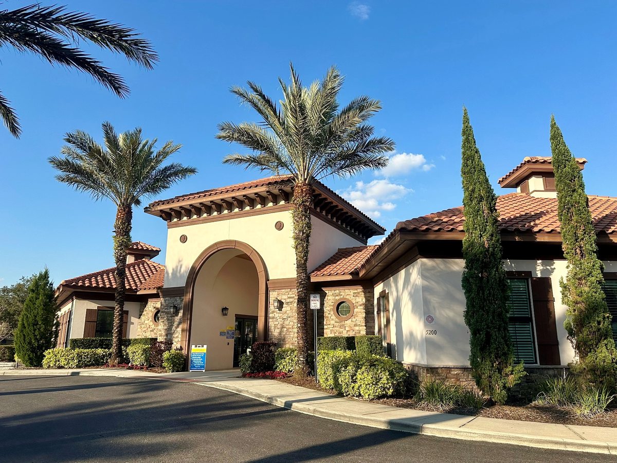 The clubhouse from an angle, showing cream stucco walls, arched doorways, a terracotta roof and slender cypress and palm trees beside a roadway.