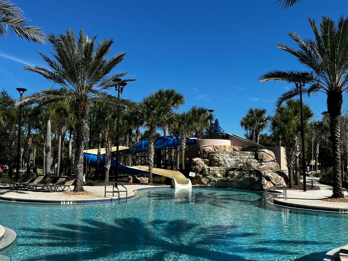 A large curving turquoise pool with a white open waterslide and a blue enclosed slide rising from a faux-rock structure, ringed by tall palms, loungers and blue parasols under a clear sky.