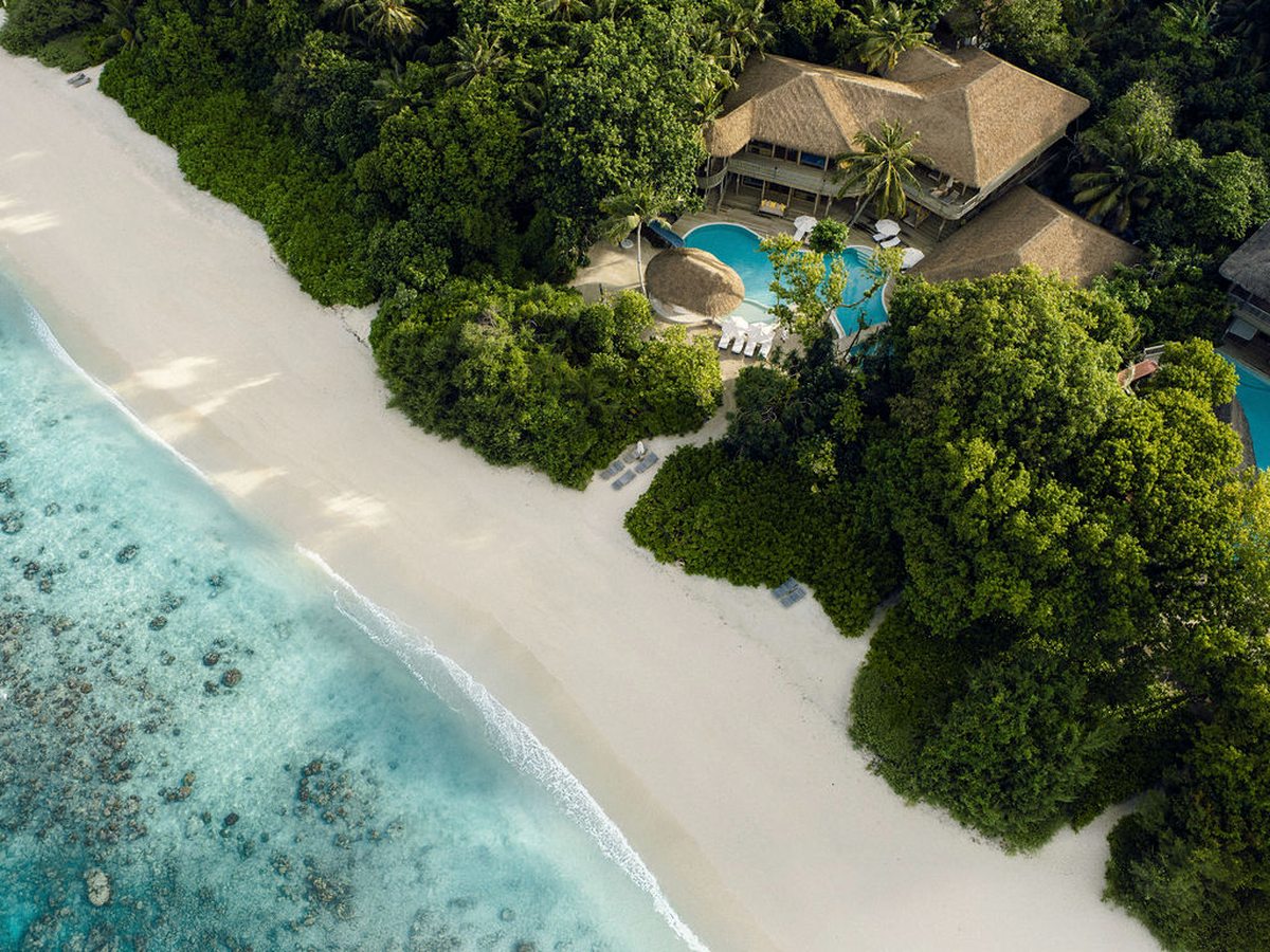 Aerial view of a thatched villa and its pool among trees beside a wide white beach, with coral visible in the clear shallows.