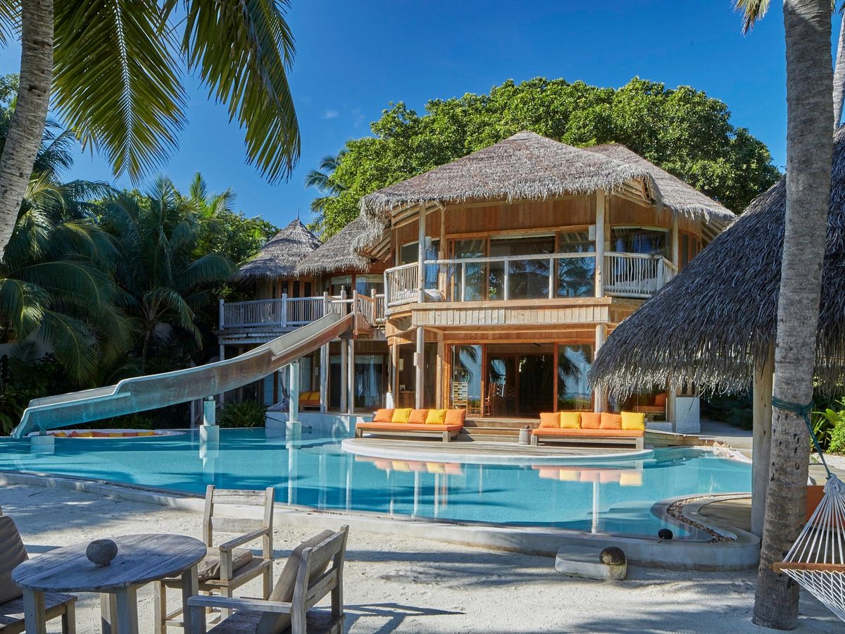 A wide blue pool in front of a two-storey thatched villa, with a white slide curving down into the water and orange daybeds along the edge.