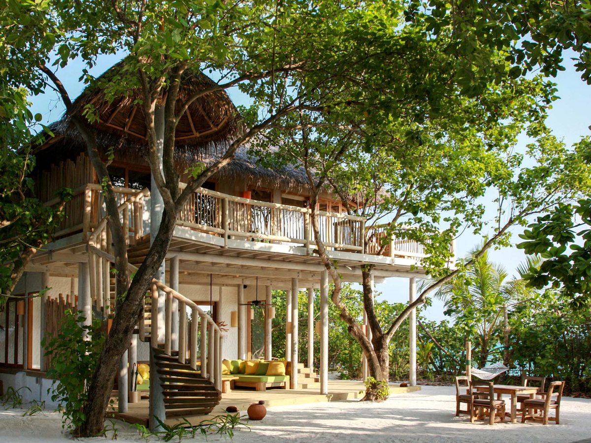 A two-storey villa with a wooden balcony and a spiral staircase standing on white sand among trees, with a table and chairs outside.