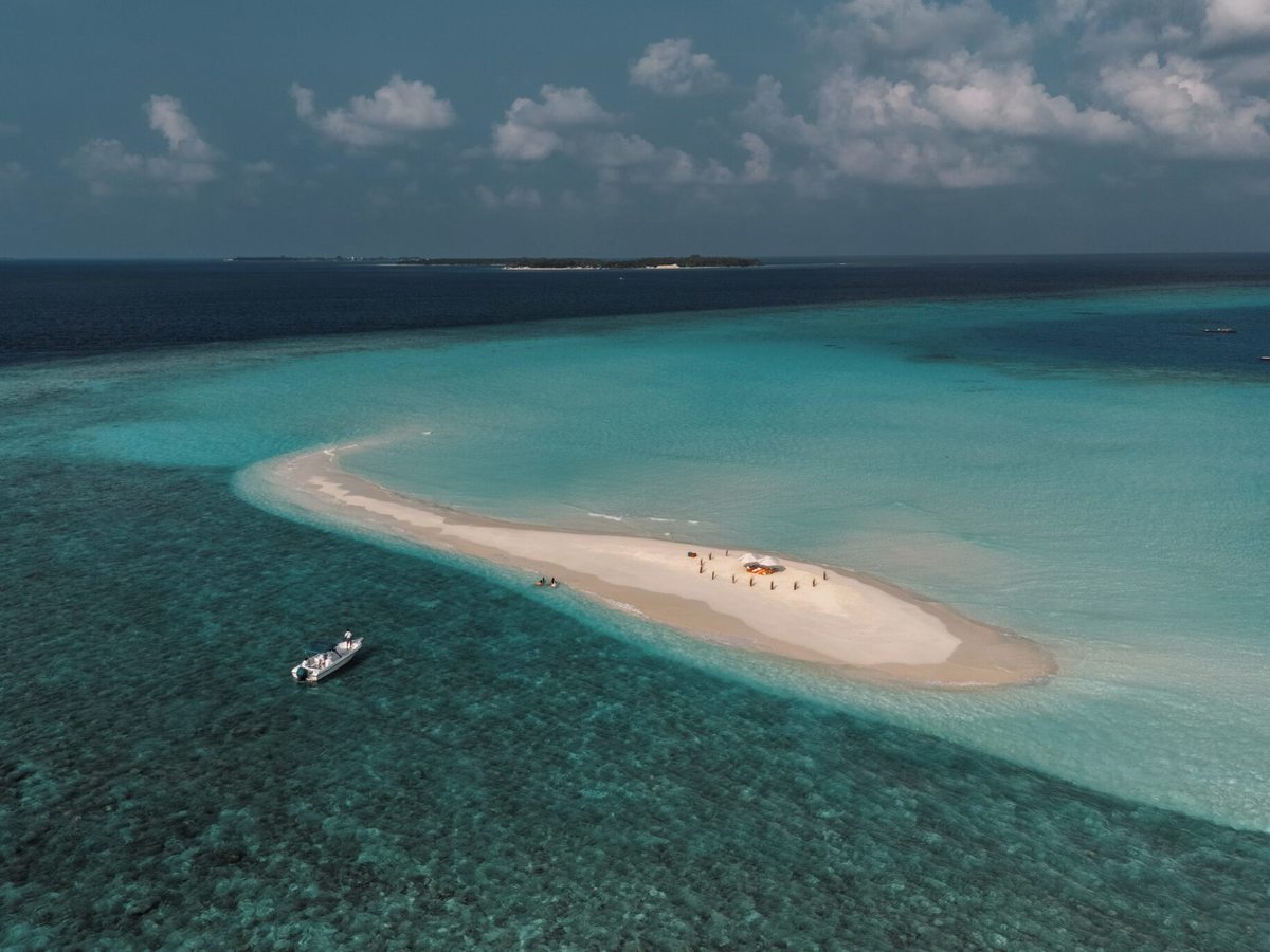 Aerial view of a long white sandbank in a turquoise lagoon with a small boat moored alongside and a shade canopy on the sand.