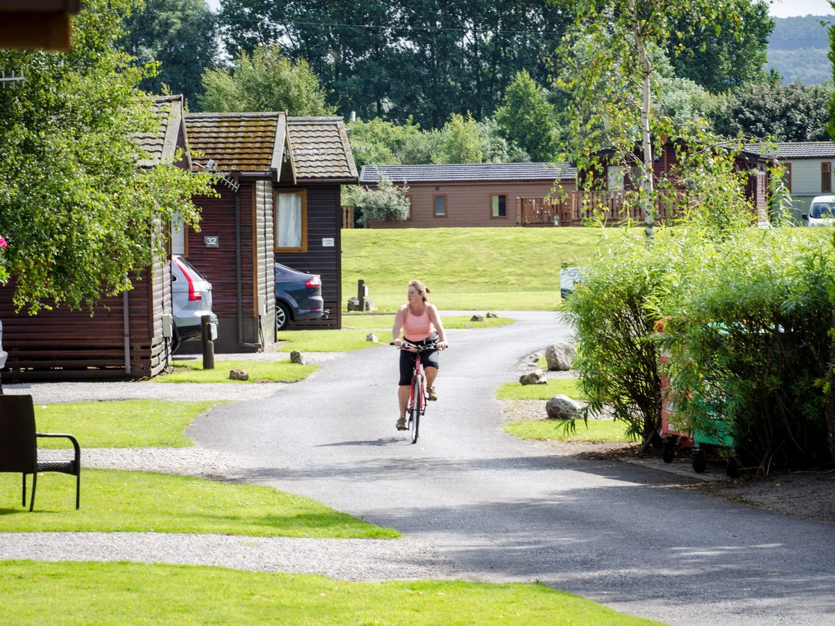 A woman cycling along a tarmac lane between timber lodges, with mown grass verges, shrubs and parked cars alongside.