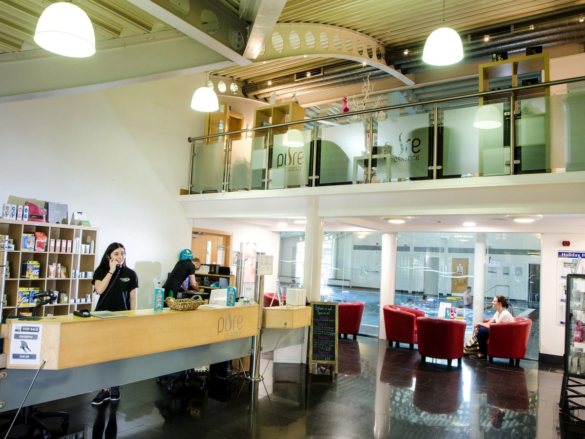 An indoor reception desk staffed by two people, with product shelves behind, red tub chairs beside a glazed wall onto the swimming pool and a mezzanine above.