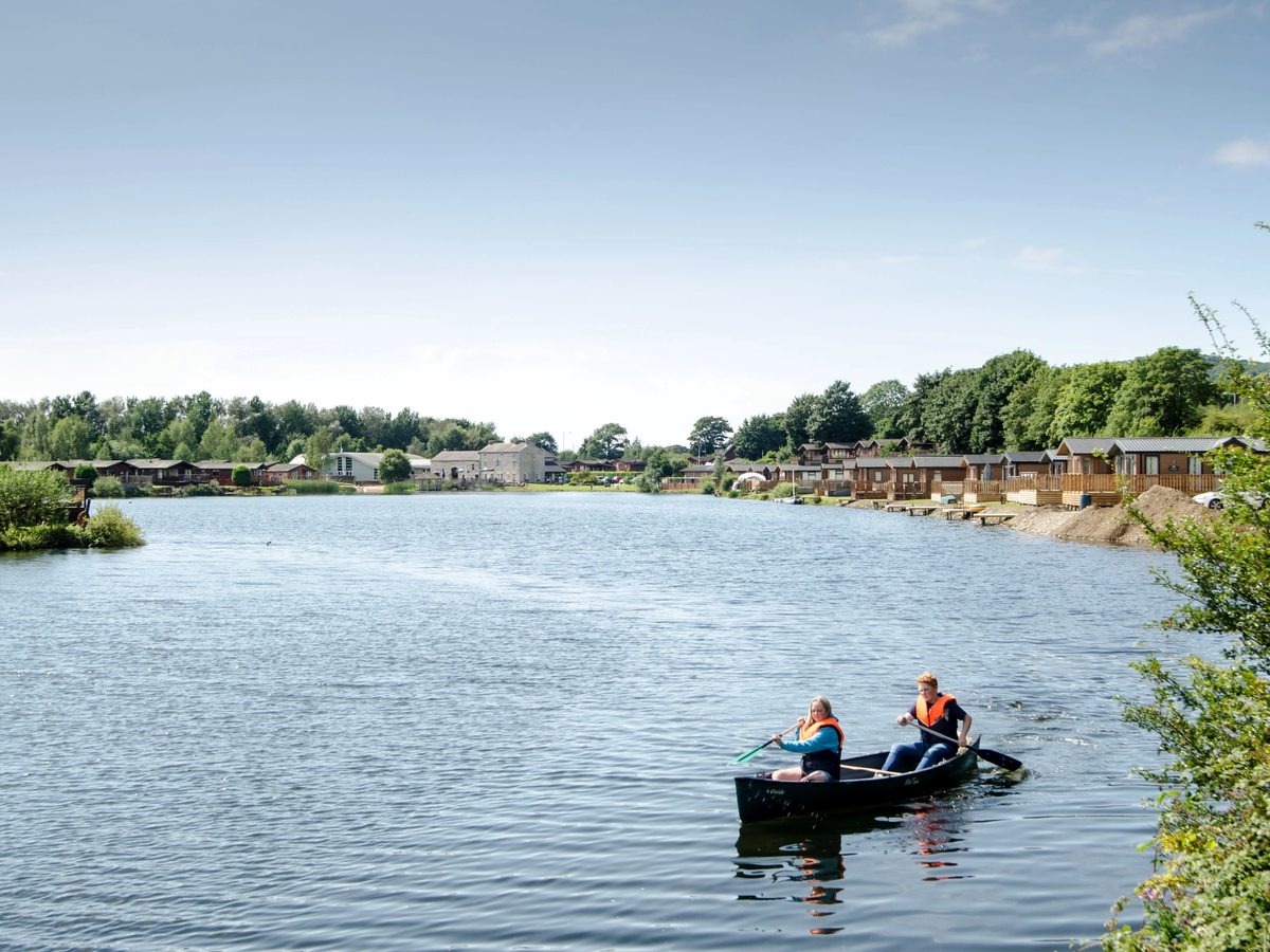 Two people paddling a canoe on a wide lake in buoyancy aids, with a wooded shoreline and a row of lodges along the far bank.