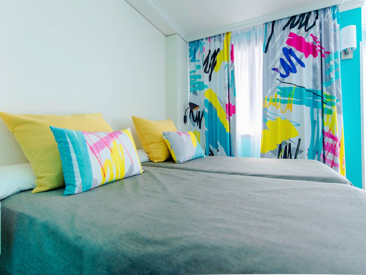 Hotel bedroom with a double bed under a grey cover with yellow and turquoise cushions, a turquoise feature wall and boldly patterned curtains at the window.
