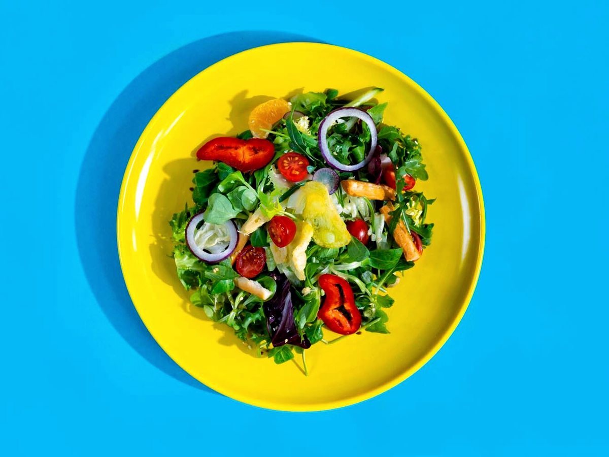 A green salad with cherry tomatoes, red onion, olives and citrus segments on a yellow plate on a turquoise table.