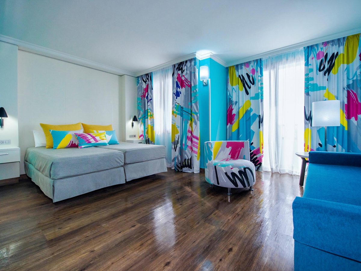 Hotel room with two joined single beds, a patterned armchair, a blue sofa and floor-length graffiti-print curtains at the balcony doors.