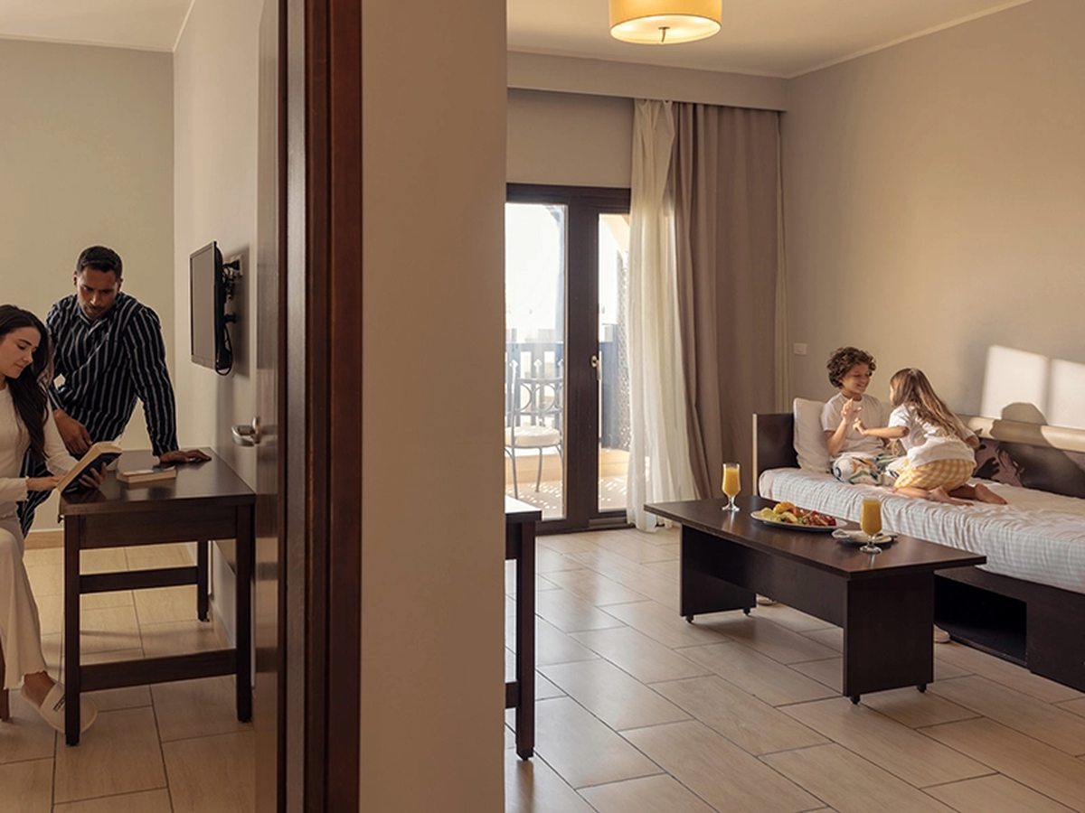 A suite living room with a sofa, coffee table and dining table, and a bedroom visible through an open doorway.