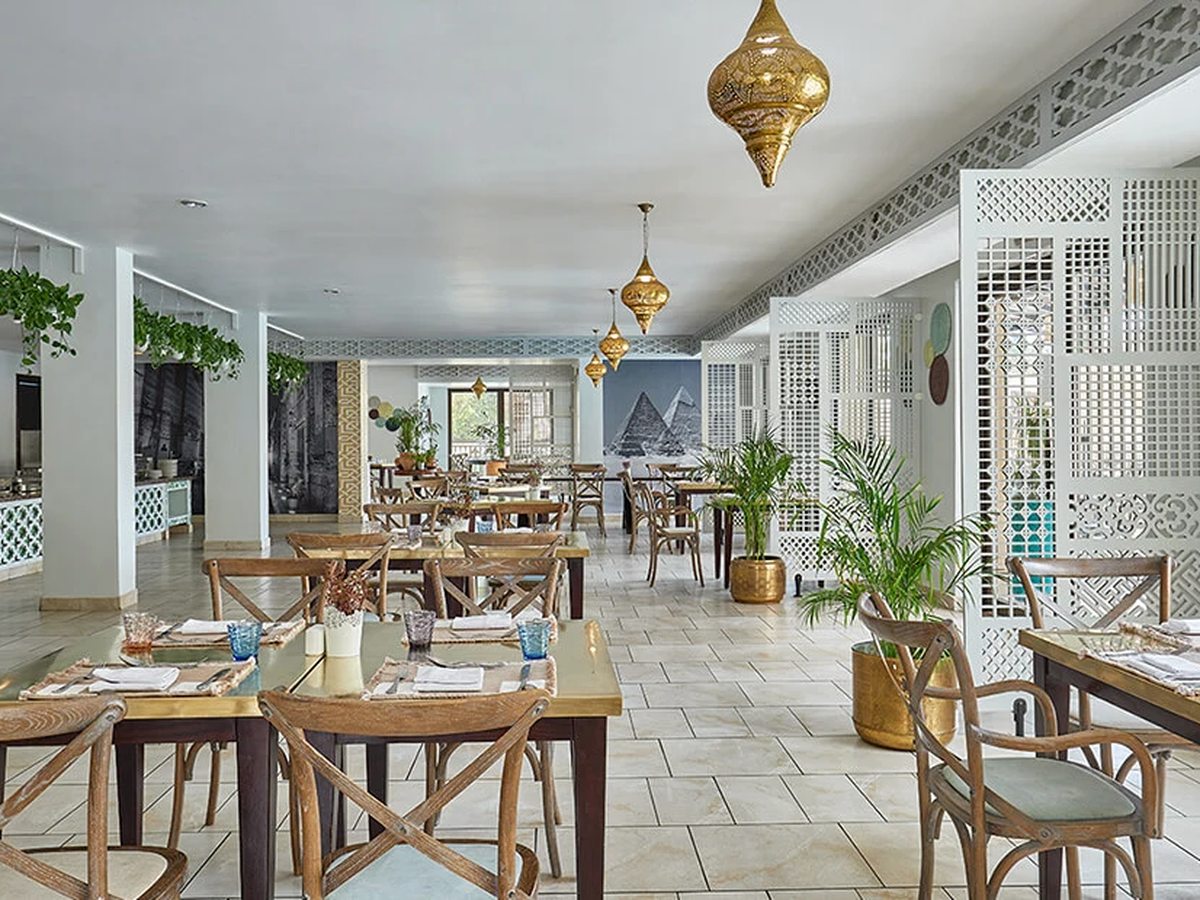 A white restaurant interior with wooden tables and chairs, gold lanterns hanging from the ceiling, fretwork screens and a mural of the pyramids.