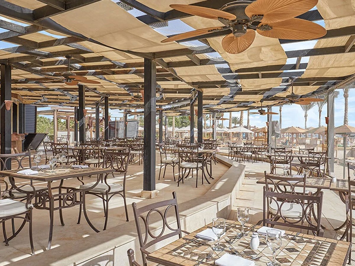 A wide covered restaurant terrace with shade sails, palm-leaf ceiling fans and mosaic-topped tables, with thatched beach parasols beyond.