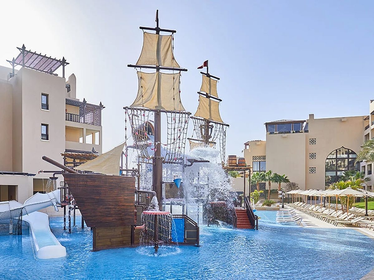 A wooden pirate ship play structure with sails and climbing nets standing in a shallow pool, water tipping from a bucket, with a small white slide alongside.