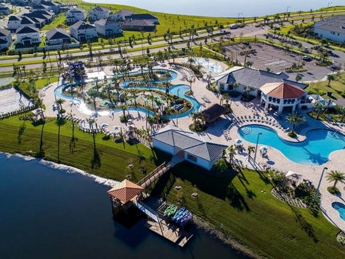Aerial across a lake with a small dock in the foreground leading to a blue pool complex and a grey-roofed clubhouse backed by trees.