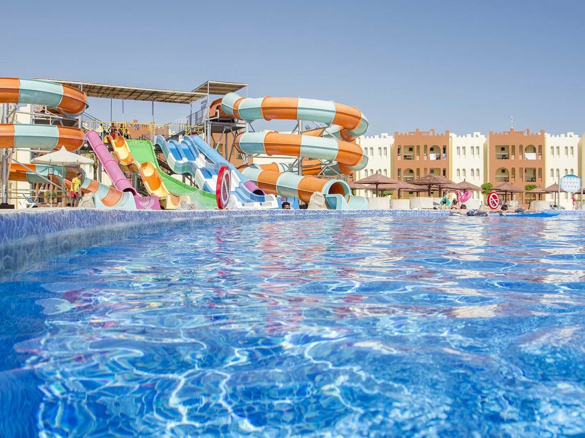 A view across a pool to a bank of orange, green, blue and pink waterslides, with cream resort blocks and thatched parasols behind.