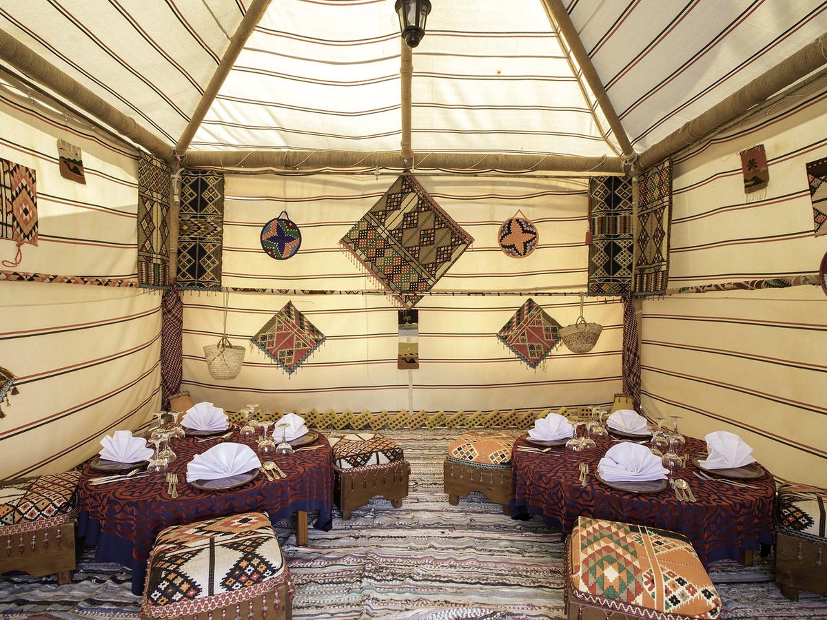 The inside of a striped Bedouin tent hung with woven textiles, with low cushioned stools around tables laid with white napkins.