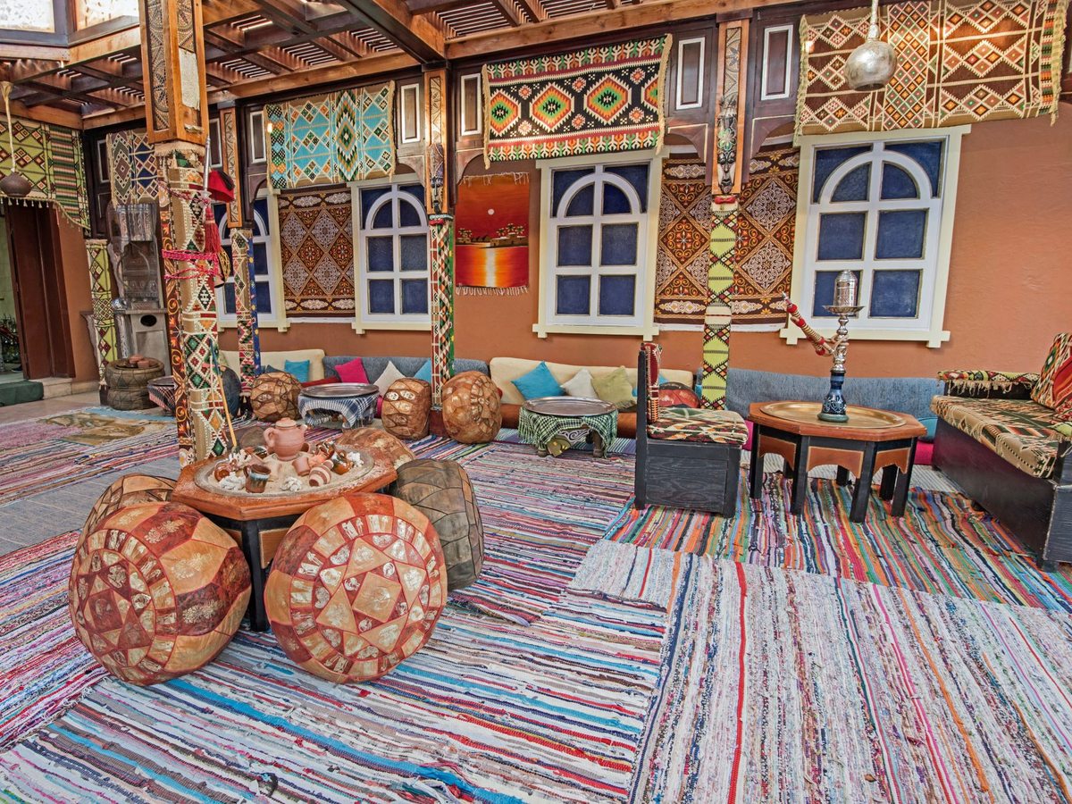 A Bedouin-style lounge with striped rag rugs, painted round stools, shisha pipes and cushioned benches under carved and painted wooden panels.