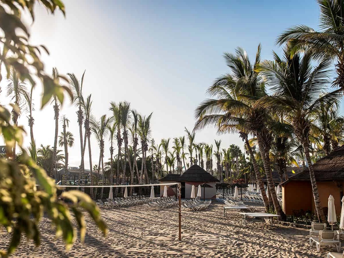Sandy artificial beach with a volleyball net strung across it, rows of sun loungers and folded parasols, thatched huts and tall palms behind.