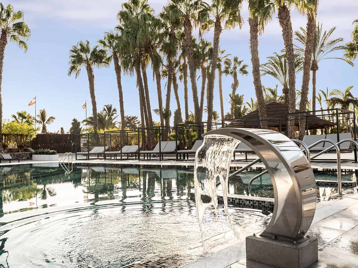 Curved chrome water cascade pouring into the shallow end of a pool, with grey sun loungers, timber daybed frames and tall palms behind.