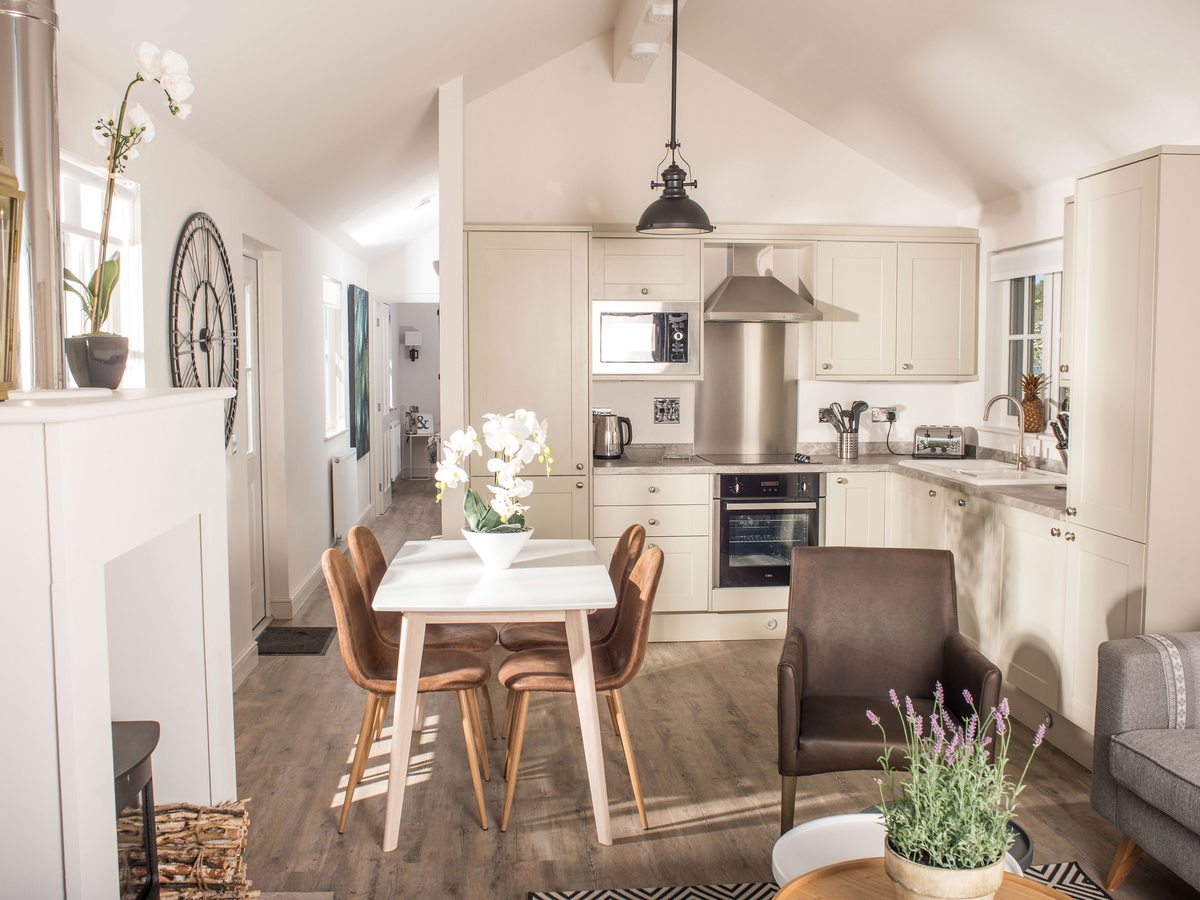 Cream fitted kitchen under a vaulted ceiling with an oven, hob, extractor hood and microwave, with a white dining table and four tan chairs in front of it.