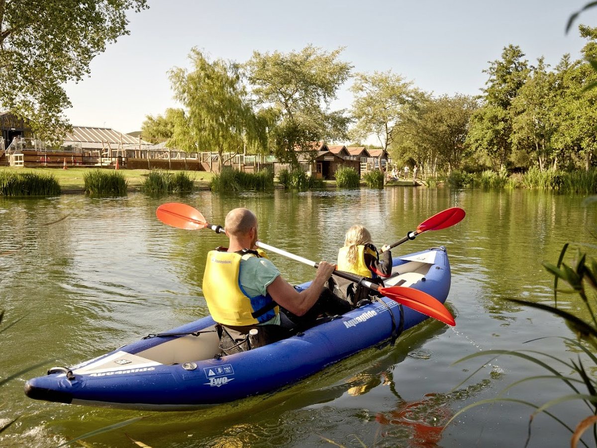 Two people in a blue tandem inflatable kayak with red paddles and buoyancy aids on the lake, with lodges and trees on the far bank.