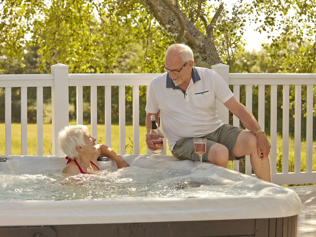 A bubbling outdoor hot tub on a raised deck with a white balustrade, two people sitting with drinks and trees behind.