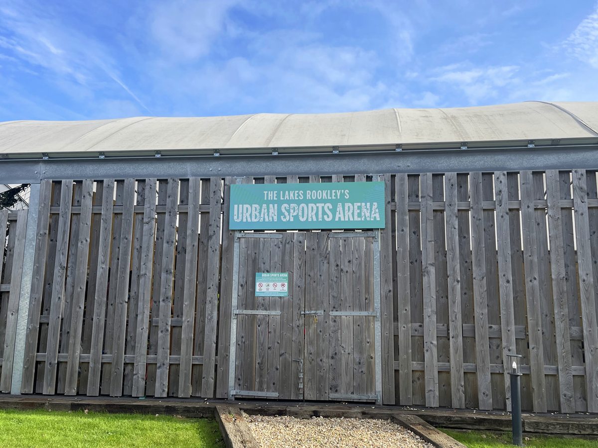 A timber-clad building with a curved fabric roof and a gated entrance signed The Lakes Rookley's Urban Sports Arena, beside a gravel path and lawn.