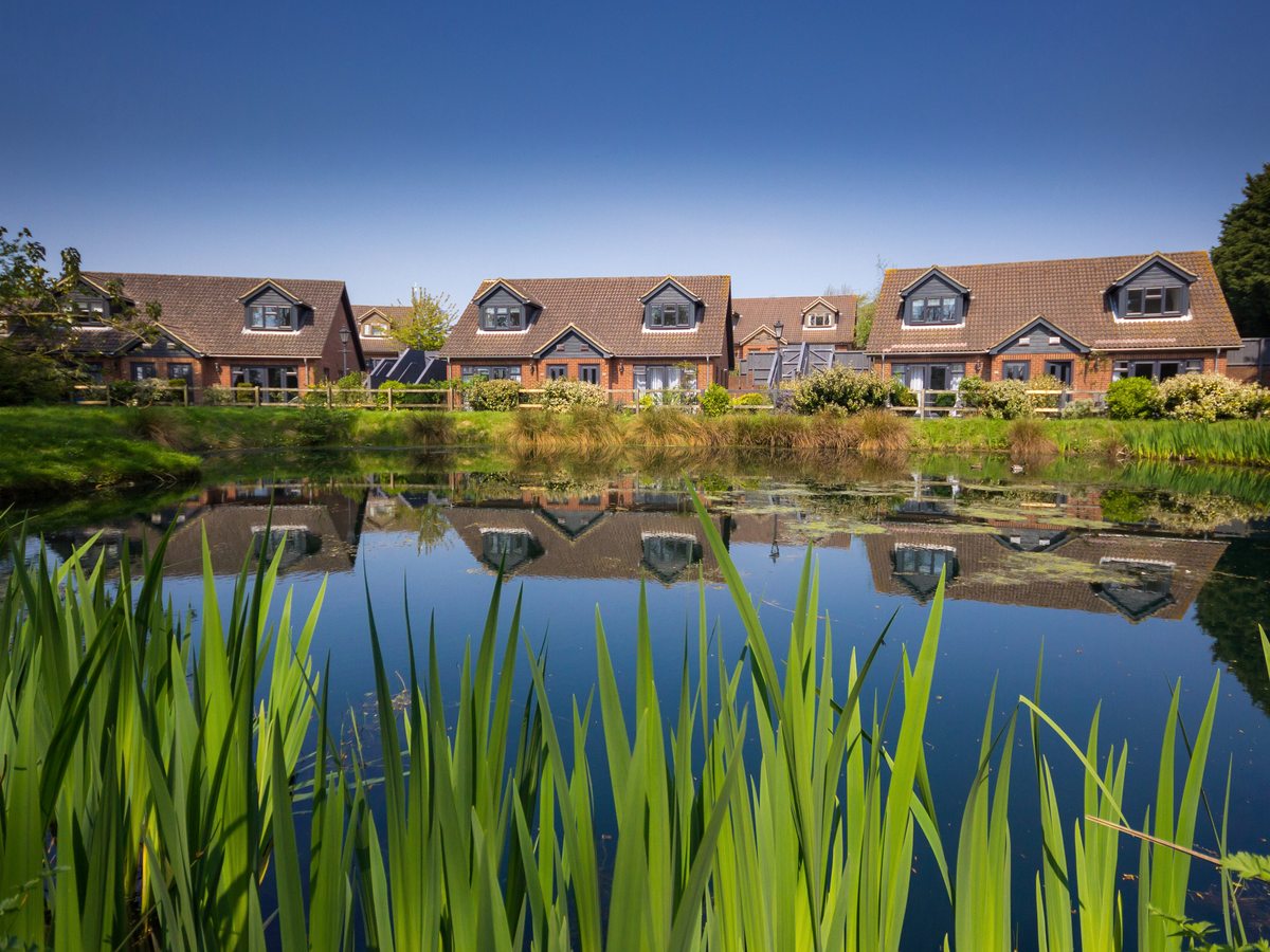 Red-brick cottages with dormer windows and tiled roofs on the bank of a lake, reflected in still water, with reeds in the foreground.