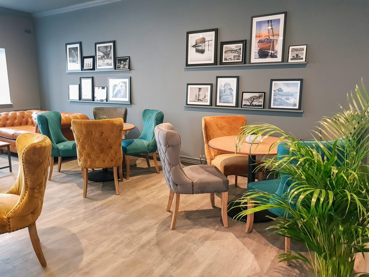 An indoor lounge with velvet armchairs in mustard, teal and grey around small round tables, a leather sofa and framed coastal photographs on a grey wall.