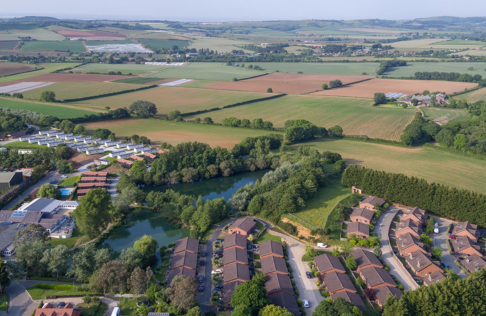Aerial view of The Lakes Rookley, with curving rows of red-roofed cottages and white caravans around two tree-fringed lakes, and the island's fields stretching away behind.