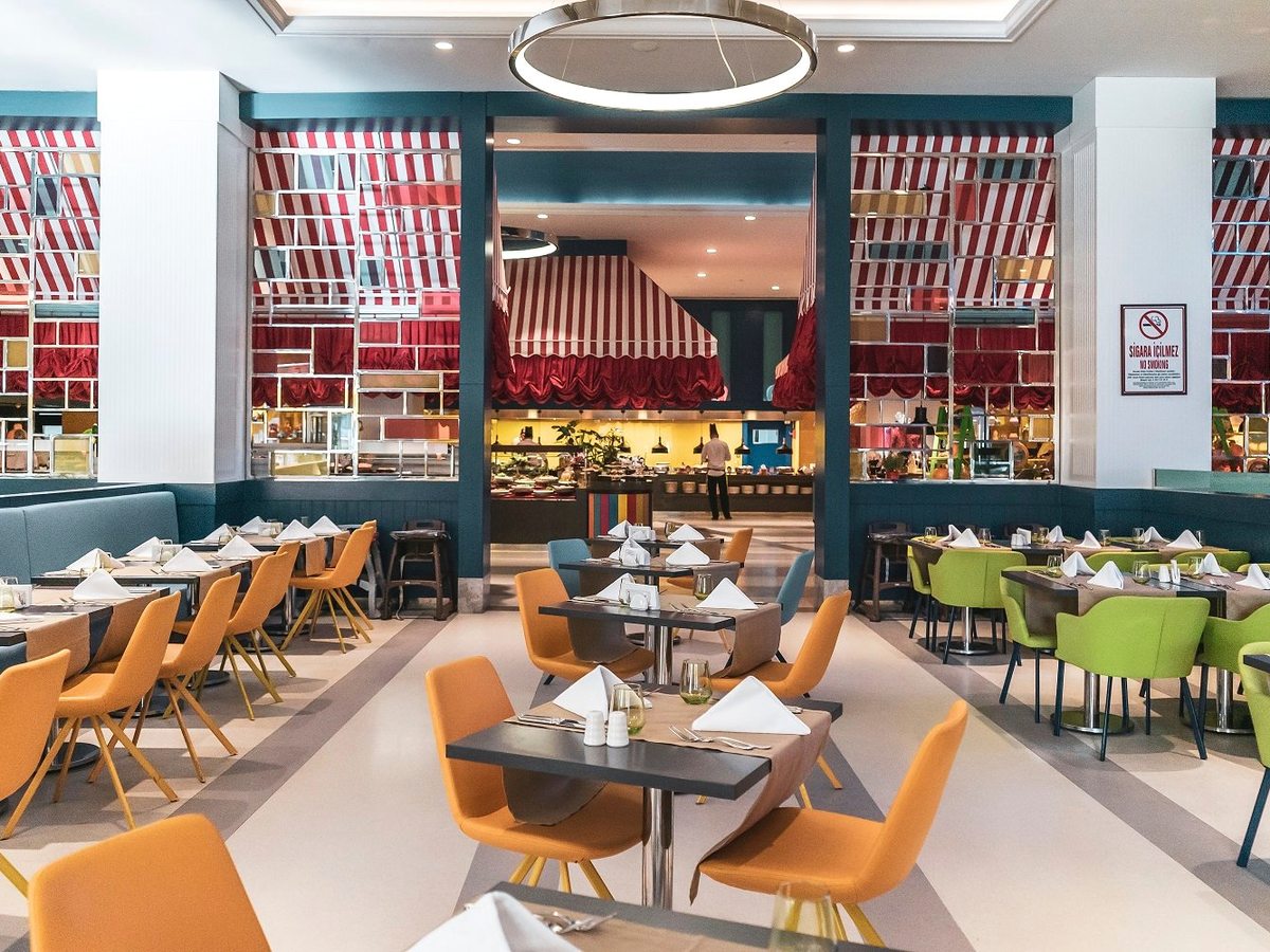 A buffet restaurant with orange and lime dining chairs at set tables, red-striped canopies over the serving counters and an open kitchen at the back.