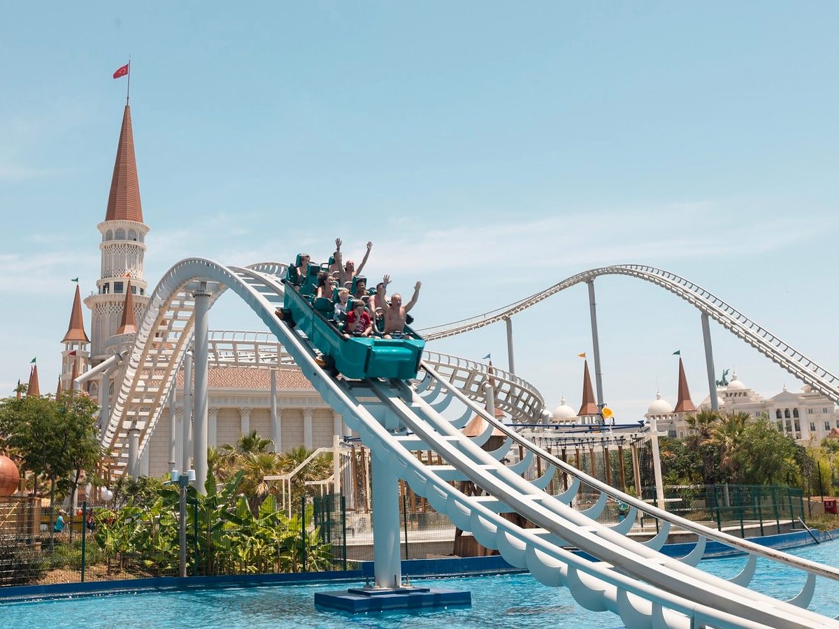 A roller-coaster train of riders with their arms raised cresting a white track above water, with the castle's spires behind.